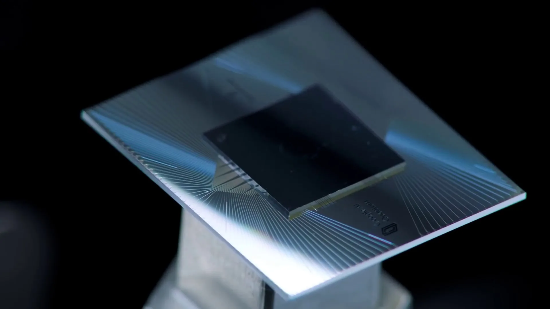 A small dark square chip mounted on a larger patterned substrate, lit from the side, with fine metal traces radiating outward