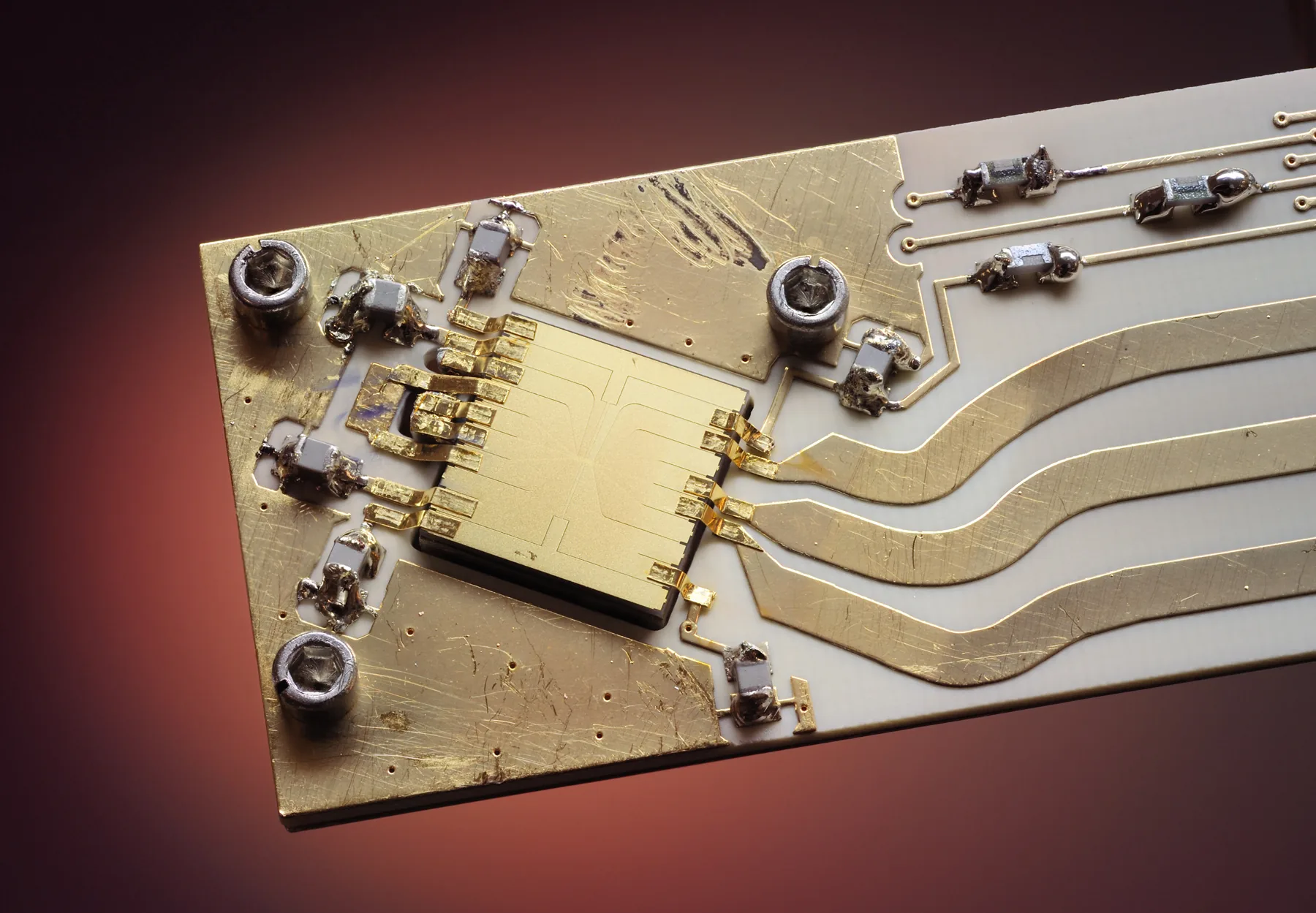A square gold ion trap mounted on a ceramic board with gold wiring and surface-mount components